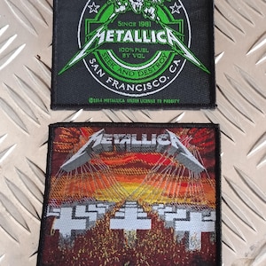Pode incluir: Dois patches da banda Metallica. O patch superior é preto com gráficos verdes e brancos, incluindo o nome da banda e "Since 1981". O patch inferior apresenta o nome da banda e "Master of Puppets" com um design de pôr do sol.