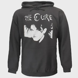 May include: A black hooded sweatshirt featuring a white graphic of a person's face and the band name "THE CURE". The sweatshirt has a front pocket and a drawstring hood. The image is a stylized portrait.