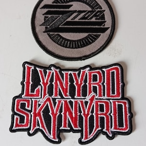 Könnte beinhalten: Zwei gestickte Aufnäher. Der obere Aufnäher ist kreisförmig mit grauem Hintergrund und einem schwarzen stilisierten "ZZ"-Design. Der untere Aufnäher ist rechteckig mit den Worten "LYNYRD SKYNRYD" in roter Schrift, weiß umrandet auf schwarzem Hintergrund.