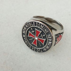 Könnte beinhalten: Ein silberfarbener Ring mit einem rot-weißem Kreuz-Design. Der Ring trägt die Inschrift "GLORIAM NON NOBIS DOMINE SED NOMINI TUO DA INC." um das Kreuz herum. Ein kleiner, klarer Stein ist in der Mitte des Kreuzes eingesetzt.