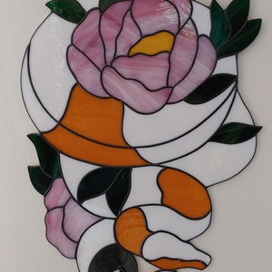 May include: A stained glass panel featuring a large pink peony with a yellow center, dark green leaves, and a white and orange snake coiled around the flower. The piece is constructed with black lead lines separating the various colored glass segments.