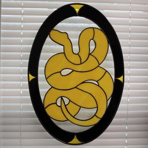 May include: A stained glass window depicting a yellow snake coiled in a circle, framed by a black oval with yellow accents.