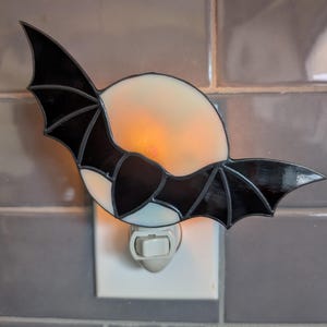 May include: A stained glass night light shaped like a bat with black wings and a cream-colored body. The night light is plugged into a white outlet and emits a warm, orange glow. The bat's wings are spread wide.