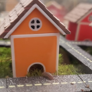 Op de afbeelding: Een klein oranje huis met een bruin dak en een rond raam. Het huis heeft een kleine opening aan de onderkant en staat op groen mos naast een miniatuurweg. Andere huizen zijn zichtbaar op de achtergrond.