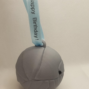 May include: A gray, spherical ornament with a textured surface, featuring a small black detail. A light blue ribbon is attached, with the words "Happy Birthday!" printed in black. The ornament is designed for gifting.