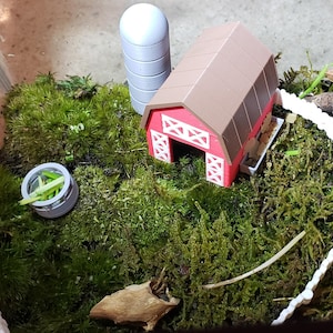 Può includere: Un fienile rosso in miniatura con tetto marrone e rivestimenti bianchi si trova su un letto di muschio verde. Una staccionata bianca circonda il fienile e un piccolo silo grigio si trova nelle vicinanze.