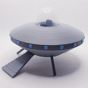 May include: A grey, 3D-printed model of a flying saucer with a clear dome and blue accents. The saucer has a ramp and three legs. The model is a novelty item.