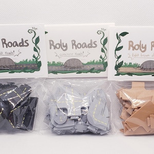 May include: Three sets of Roly Roads toy road pieces in clear plastic bags. Each set has a label with the text "Roly Roads" and a different road type: asphalt, concrete, and light brown dirt roads. Each set contains 33 pieces.