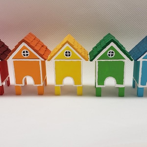 Op de afbeelding: Een rij van acht miniatuurhouten huisjes, geschilderd in verschillende kleuren, waaronder roze, rood, oranje, geel, groen, blauw, bruin en beige. Elk huisje heeft een witte deur en een wit raam.