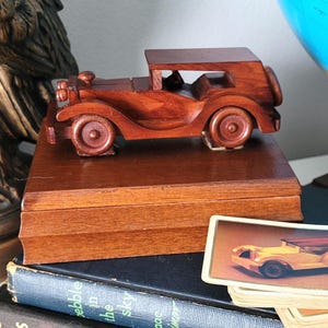 May include: A wooden model of a vintage car sits atop a wooden box, with a stack of playing cards featuring the same car design. The cards are next to a stack of books. An Ace of Spades card is visible.