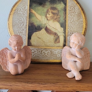 May include: A decorative plaque featuring a central rectangular print of a child pointing, framed by a gold and cream border with scrollwork, flanked by two terracotta-colored angel figurines, one kneeling with hands clasped and one sitting with a hand on its cheek.