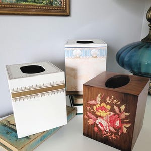 May include: Three decorative tissue box covers. One is white with gold trim, another has a seashell design, and the third is wood with a floral design. The boxes are square with a circular opening on top.