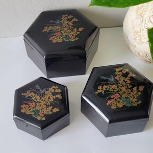 May include: Three black hexagonal boxes with glossy finishes, each featuring a decorative floral motif on the lid. The design displays clusters of orange, yellow, and green flowers with thin, dark stems, arranged in a consistent pattern across all lids.