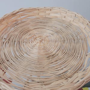 May include: A two-tiered circular basket stand constructed from light tan woven wicker. The design features two round, shallow trays connected by a central vertical support, showcasing a natural, textured, and interlaced reed pattern throughout.