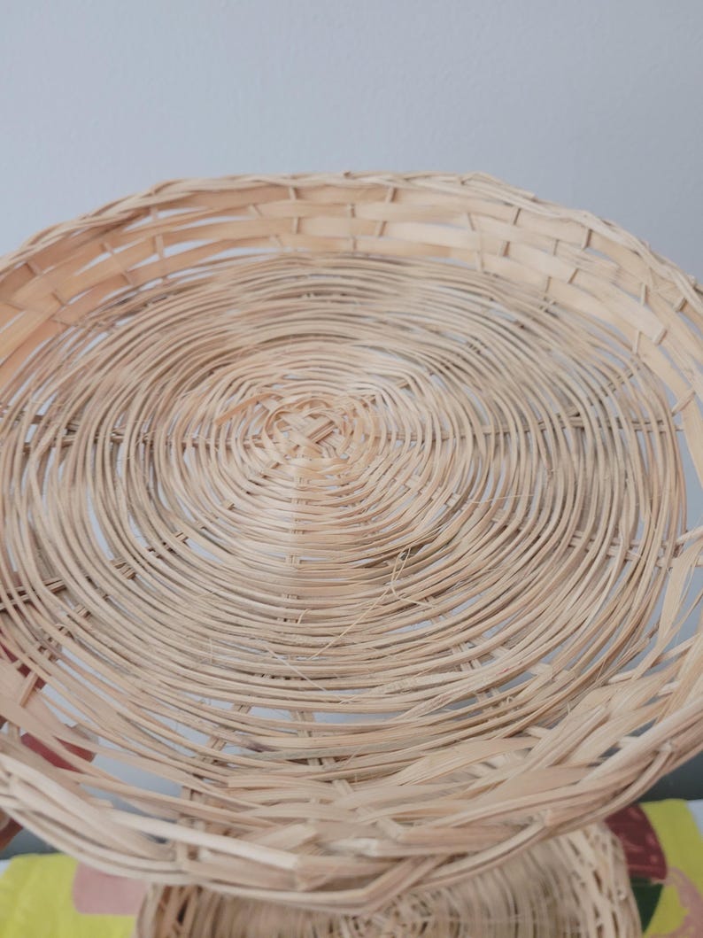 May include: A two-tiered circular basket stand constructed from light tan woven wicker. The design features two round, shallow trays connected by a central vertical support, showcasing a natural, textured, and interlaced reed pattern throughout.