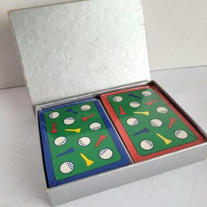 May include: A silver playing card box with two decks of cards. The cards feature a green background with golf balls and tees. One deck has a blue border, the other a red border. The box reads "Fields & Meyer Playing Cards."