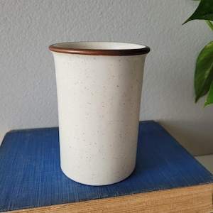 May include: A cylindrical ceramic mug with a speckled off-white finish and a smooth, dark brown rim. The mug features a straight-sided design with a flat base, showcasing a simple, unadorned aesthetic without a handle or any additional decorative elements.