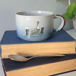 May include: A light blue ceramic mug with a speckled finish and a brown-rimmed handle. The mug features a graphic illustration of a pelican standing on a wooden dock. The mug is positioned on top of two stacked hardcover books with a silver spoon.
