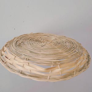 May include: A shallow, round, hand-woven basket made from light-colored, natural dried reeds. The basket features a tightly coiled, circular base that transitions into a wider, open-weave rim with vertical supports, creating a flat, bowl-like shape.