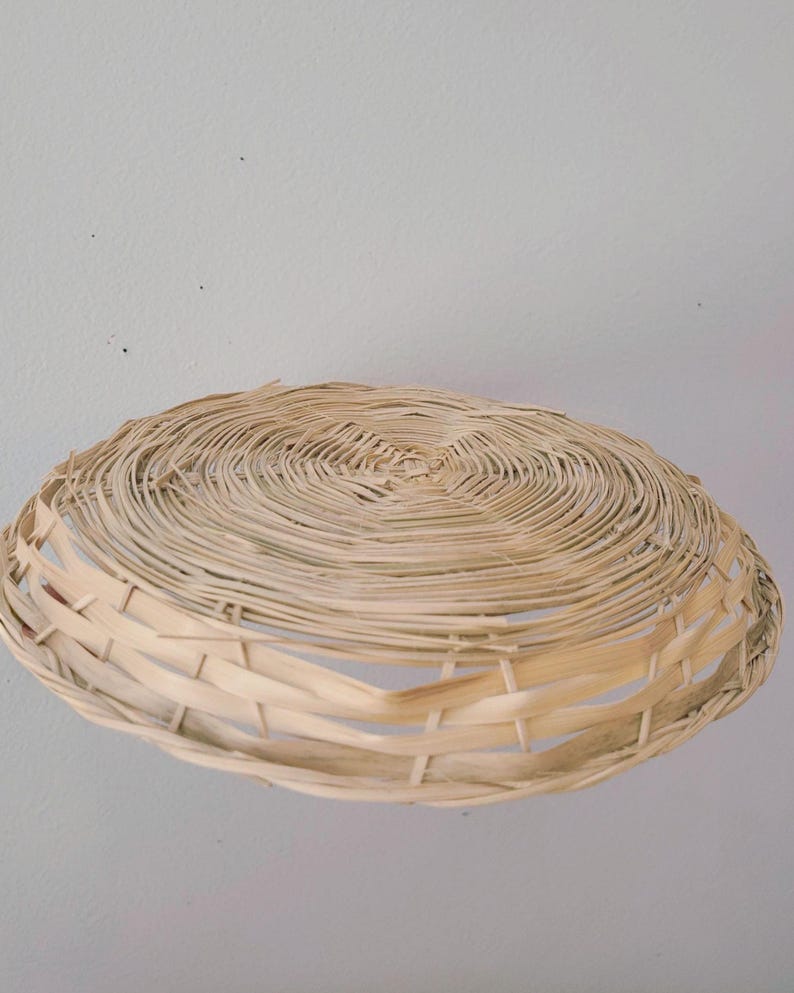 May include: A shallow, round, hand-woven basket made from light-colored, natural dried reeds. The basket features a tightly coiled, circular base that transitions into a wider, open-weave rim with vertical supports, creating a flat, bowl-like shape.