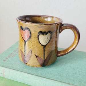 May include: A ceramic mug with a glossy mustard-yellow glaze featuring two hand-painted tulip designs. One tulip is colored in soft orange and the other in cream, both with dark brown stems and leaves. The mug has a dark brown rim and handle.