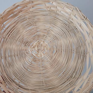 May include: A round, shallow basket woven from light-colored, natural reed strips. The design features a concentric circular pattern radiating from the center, with thin, flexible strands interlaced to form a sturdy, open-weave structure.