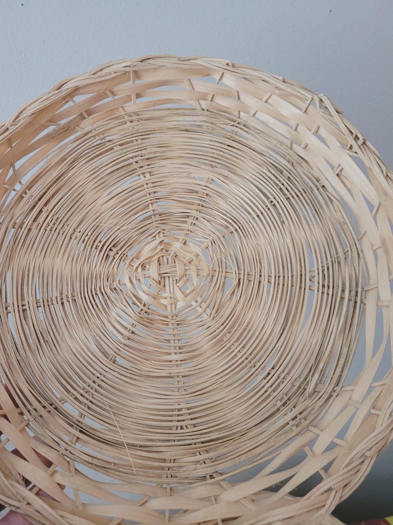 May include: A round, shallow basket woven from light-colored, natural reed strips. The design features a concentric circular pattern radiating from the center, with thin, flexible strands interlaced to form a sturdy, open-weave structure.