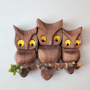 May include: A wall-mounted wooden carving featuring three owls with textured brown bodies and bright yellow circular eyes. The owls are perched together on a single horizontal branch that includes a small green leaf accent on the left side.