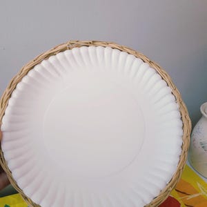 May include: A round white plate with a fluted edge, securely nested within a circular woven wicker basket holder. The plate features a smooth, flat center and a textured, scalloped rim, while the basket is crafted from light brown natural fibers.
