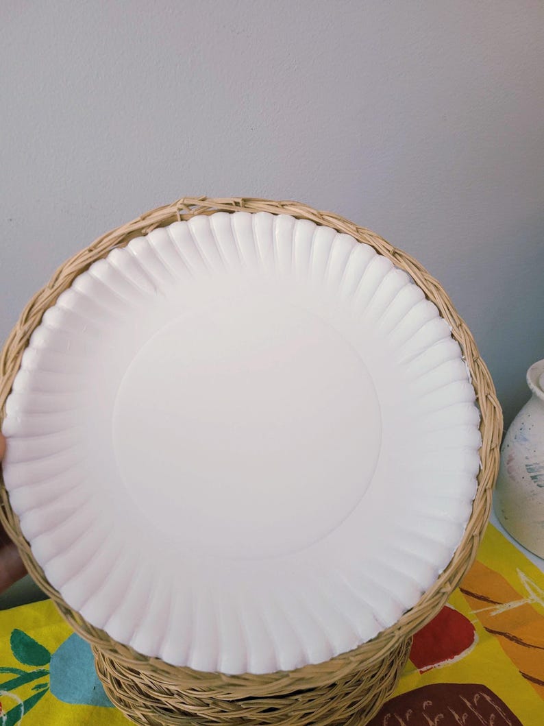 May include: A round white plate with a fluted edge, securely nested within a circular woven wicker basket holder. The plate features a smooth, flat center and a textured, scalloped rim, while the basket is crafted from light brown natural fibers.