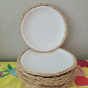 May include: A stack of round white paper plates, each fitted into a circular light brown woven wicker holder. The holders feature a braided rim design that surrounds the edge of the plates, creating a layered appearance for the set of dining items.