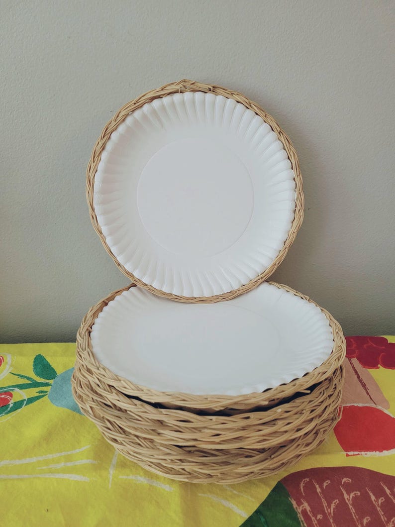 May include: A stack of round white paper plates, each fitted into a circular light brown woven wicker holder. The holders feature a braided rim design that surrounds the edge of the plates, creating a layered appearance for the set of dining items.