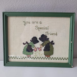 May include: A framed cross-stitch embroidery featuring two figures in dark bonnets and green dresses holding pink flowers. The text reads, "you are a Special Friend." The piece is enclosed in a rectangular green wooden frame with a checkered border.
