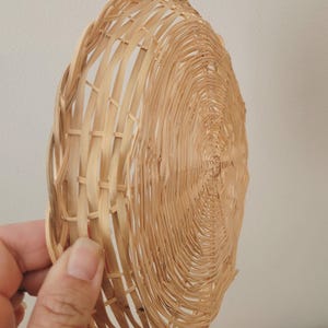 May include: A round, shallow basket crafted from light tan woven reeds. The design features a spiral pattern radiating from the center, with a structured, open-weave border along the edge, showcasing the natural texture and color of the material.