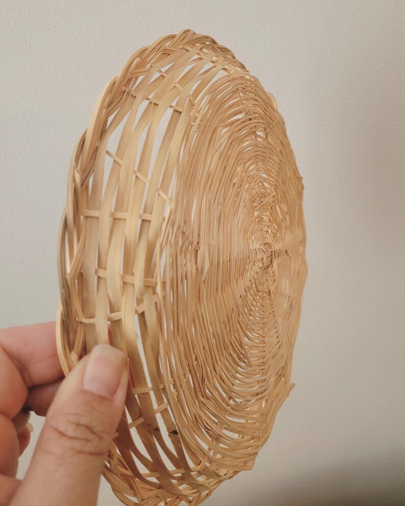 May include: A round, shallow basket crafted from light tan woven reeds. The design features a spiral pattern radiating from the center, with a structured, open-weave border along the edge, showcasing the natural texture and color of the material.