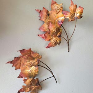 May include: Two wall-mounted metal leaf sculptures featuring a metallic copper and bronze finish with iridescent pink and orange highlights. Each piece consists of three interconnected maple-shaped leaves with textured surfaces and thin stems.