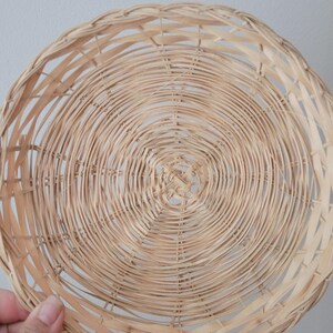 May include: A round, shallow basket crafted from light-colored, natural woven wicker material. The design features a concentric circular pattern radiating from the center, with thin, flexible strips of wood interlaced to form a sturdy, open-weave structure.
