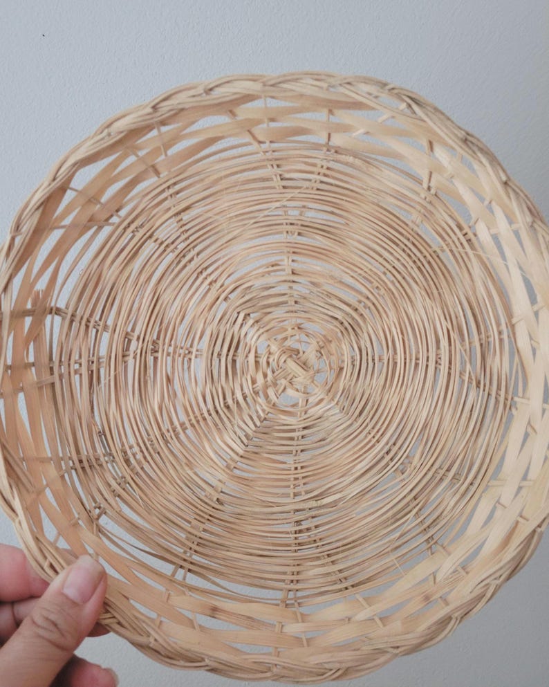 May include: A round, shallow basket crafted from light-colored, natural woven wicker material. The design features a concentric circular pattern radiating from the center, with thin, flexible strips of wood interlaced to form a sturdy, open-weave structure.