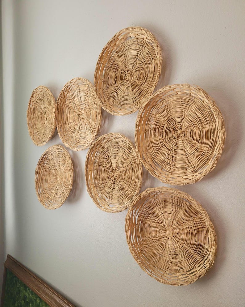 May include: A collection of six circular woven wicker wall baskets in varying sizes, featuring a natural light tan color and a concentric spiral pattern. The baskets are arranged in a cluster, each displaying a distinct woven texture and rim.