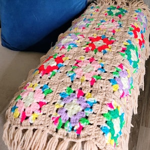 May include: A handmade crochet blanket featuring a series of square motifs joined together. Each square has a beige border with a colorful center in shades of pink, red, green, yellow, and purple. The blanket is finished with a long beige fringe.