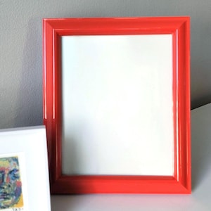 May include: A rectangular picture frame with a glossy, vibrant red finish and a smooth, beveled edge design. The frame features a clean, white interior space, providing a stark contrast to the bold red border of the frame structure.