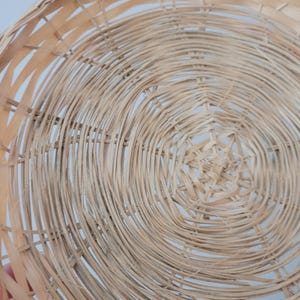 May include: A round, shallow basket crafted from light-colored, natural woven reeds. The design features a concentric circular pattern radiating from the center, with thin, flexible strips of wood interlaced to form a sturdy, open-weave structure.