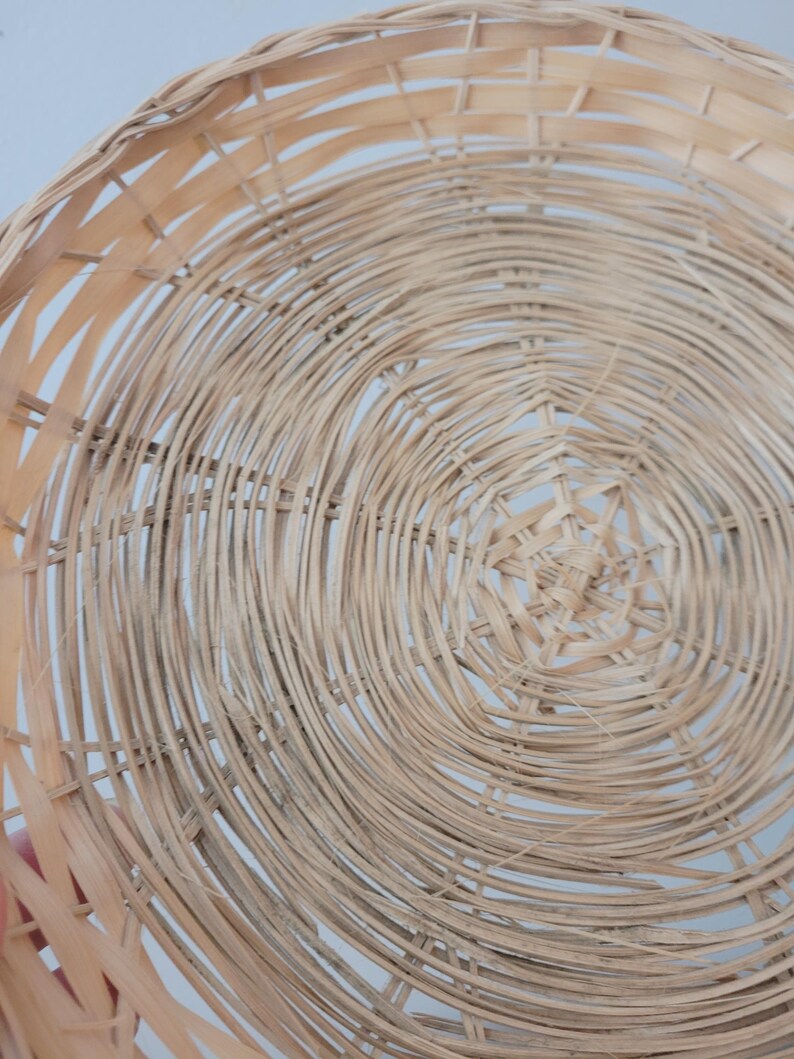 May include: A round, shallow basket crafted from light-colored, natural woven reeds. The design features a concentric circular pattern radiating from the center, with thin, flexible strips of wood interlaced to form a sturdy, open-weave structure.