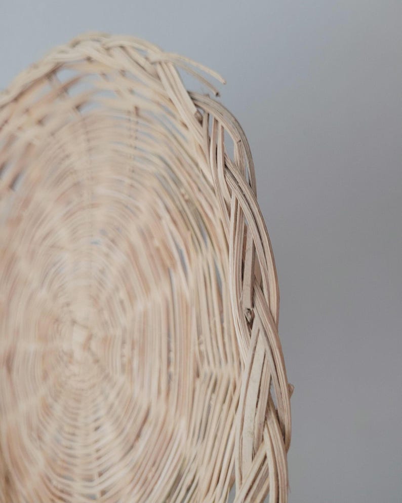 May include: A round, light beige woven wicker tray featuring a concentric circular pattern and a braided edge. The natural material displays a textured, interlaced design with visible fibers, creating a shallow, flat, and circular structure.