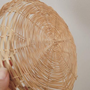 May include: A round, shallow basket woven from light tan natural reeds. The structure features a circular base with radiating spokes and a continuous spiral weave pattern that extends outward to form the curved, open-work sides of the object.