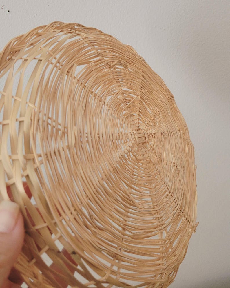 May include: A round, shallow basket woven from light tan natural reeds. The structure features a circular base with radiating spokes and a continuous spiral weave pattern that extends outward to form the curved, open-work sides of the object.