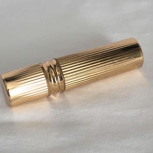 May include: A gold-colored, cylindrical perfume vial with a ribbed design. The vial has a cap on one end and a decorative band in the middle. The surface of the vial is shiny and reflects light.