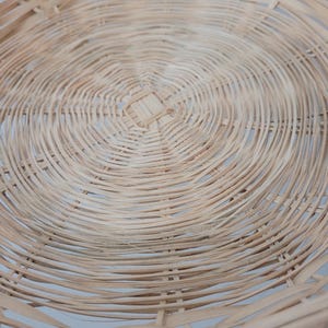 May include: A round, shallow basket woven from light tan natural reed or wicker material. The design features a concentric circular pattern radiating from a square-shaped center weave, with thin, flexible strips forming the base and sides.