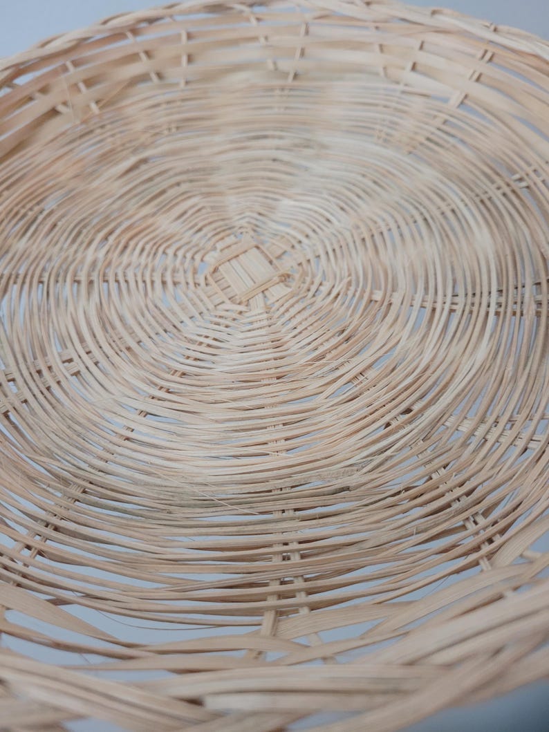 May include: A round, shallow basket woven from light tan natural reed or wicker material. The design features a concentric circular pattern radiating from a square-shaped center weave, with thin, flexible strips forming the base and sides.