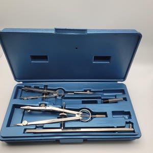 May include: A blue plastic storage case containing a set of silver metal drafting compasses and a straight metal extension bar. The interior of the case is molded to fit each tool securely, with two compasses and one bar visible in the compartments.